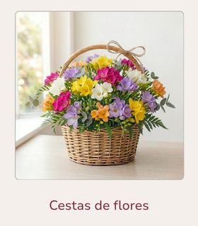 Cesta de mimbre con flores de fressia en diferentes colores. La cesta de flores está colocada encima de una mesa clara de madera.