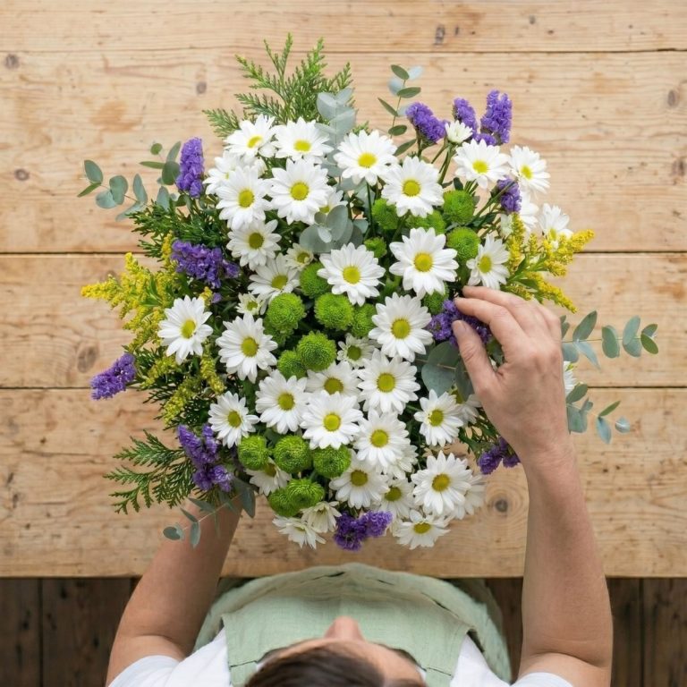 Vista cenital de un florista preparando un ramo de flores variadas en color blanco, verde y morado.